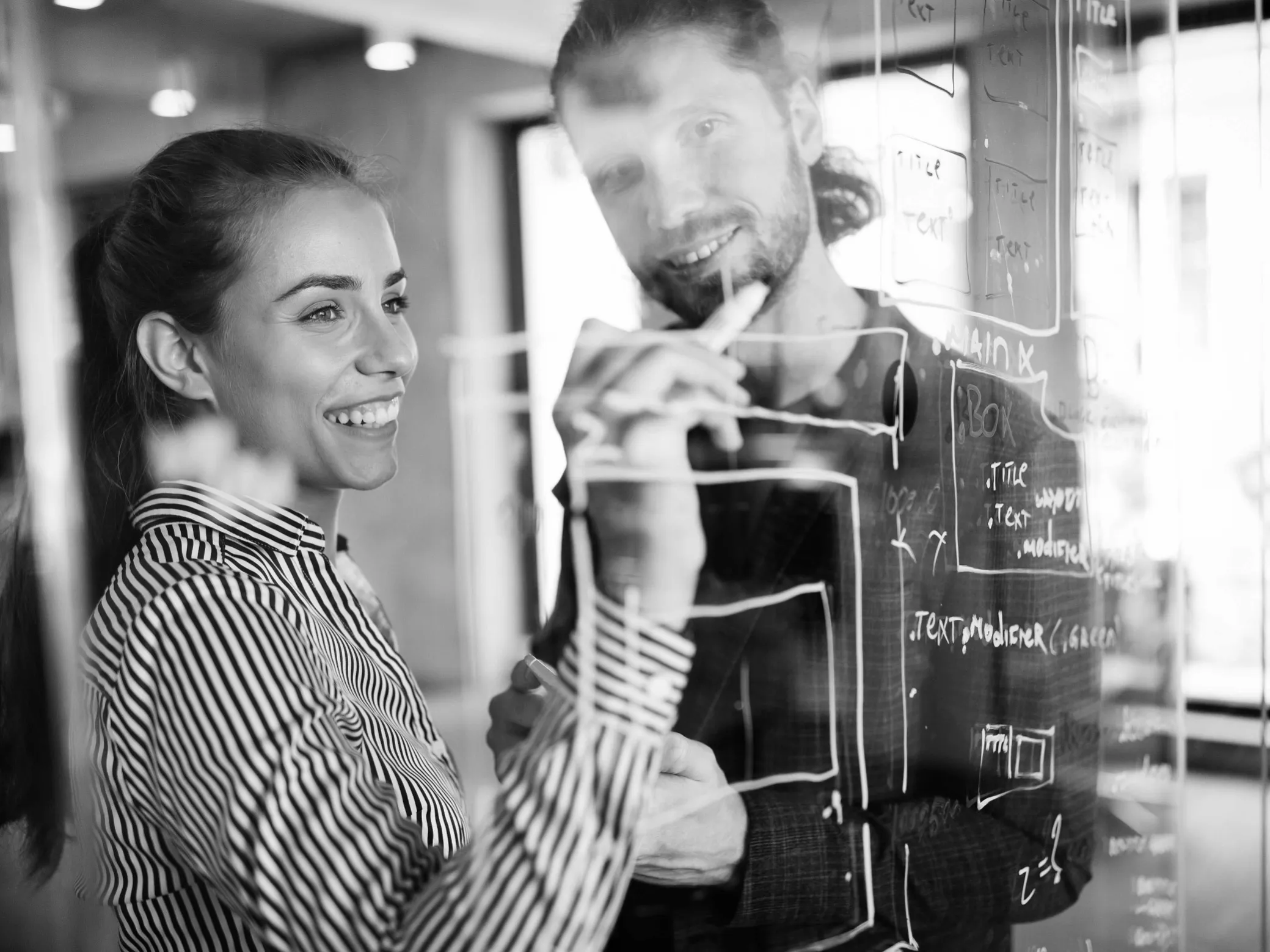 Two colleagues brainstorming and drawing diagrams on a transparent glass board in an office.
